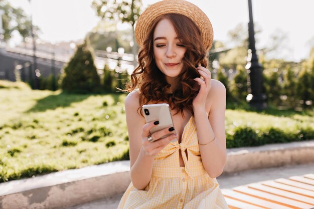 Agradable chica caucásica con mensaje de texto de cabello rojo en el parque Disparo al aire libre de juguetona mujer jengibre con sombrero usando su teléfono en el fondo de la naturaleza