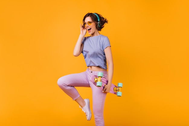 Agradable chica caucásica en pantalones pitillo rosas bailando sobre fondo naranja Foto de estudio de dama rizada en auriculares posando con longboard y riendo