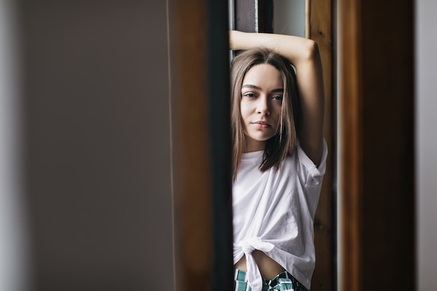 Agradable chica europea con el pelo lacio de pie en su piso y mirando. Encantadora mujer caucásica morena posando con las manos en alto.