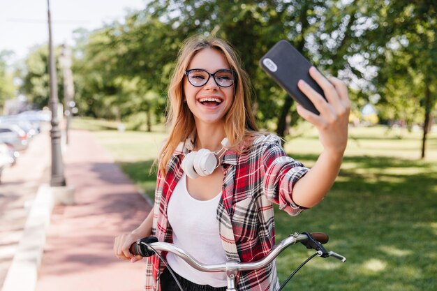 Agradable chica rubia con smartphone para selfie en parque. Encantadora dama sonriente con gafas montando en bicicleta.
