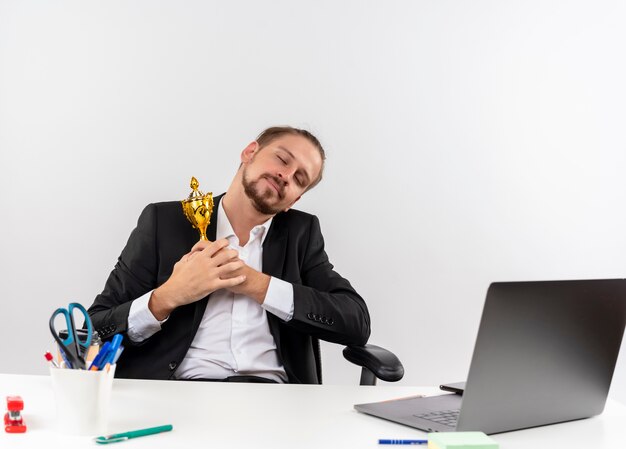 Agradable empresario guapo en traje sosteniendo trofeo sentado en la mesa en offise sobre fondo blanco.