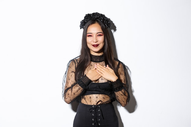 Agradable hermosa mujer asiática en traje de bruja sonriendo, tomados de la mano en el pecho y mirando agradecida, expresa gratitud, de pie en traje de halloween sobre fondo blanco.