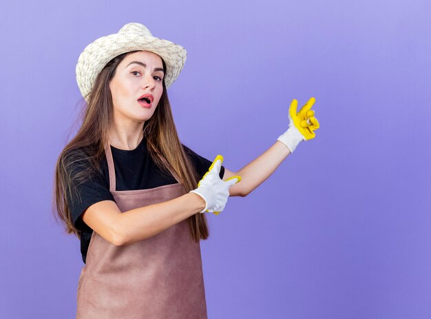 Agradable jardinero hermosa chica en uniforme con sombrero y guantes de jardinería puntos en la parte posterior aislado en azul