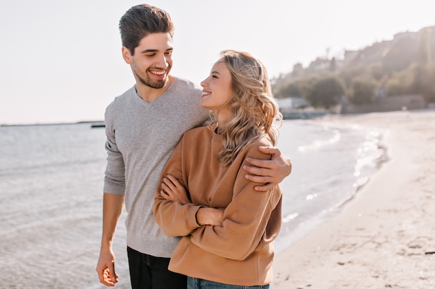 Agradable joven abrazando a novia en la naturaleza. Retrato al aire libre de niña rubia complacida posando en el mar con su marido.