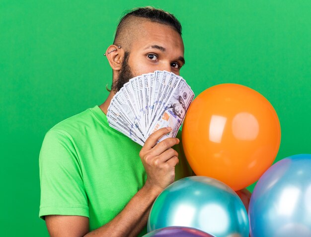 Agradable joven afroamericano vistiendo camiseta verde de pie detrás de globos con la cara cubierta con dinero en efectivo