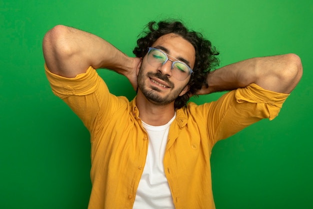 Agradable joven apuesto hombre caucásico con gafas poniendo las manos detrás de la cabeza con los ojos cerrados aislado sobre fondo verde