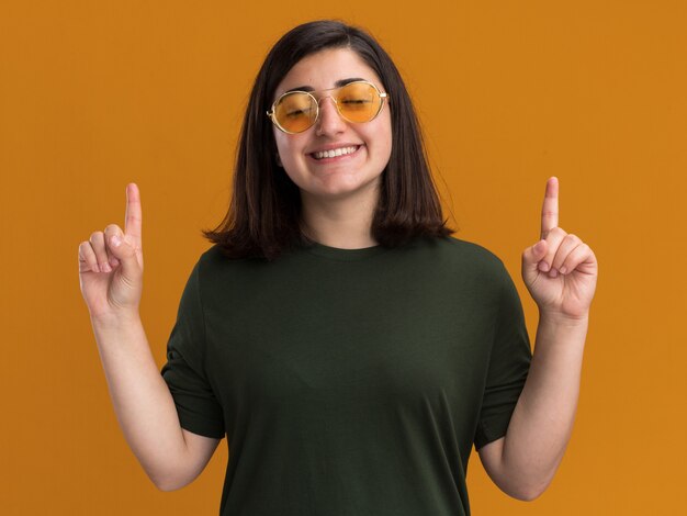 Agradable joven bastante caucásica en gafas de sol se encuentra con los ojos cerrados y apunta hacia arriba aislado en la pared naranja con espacio de copia