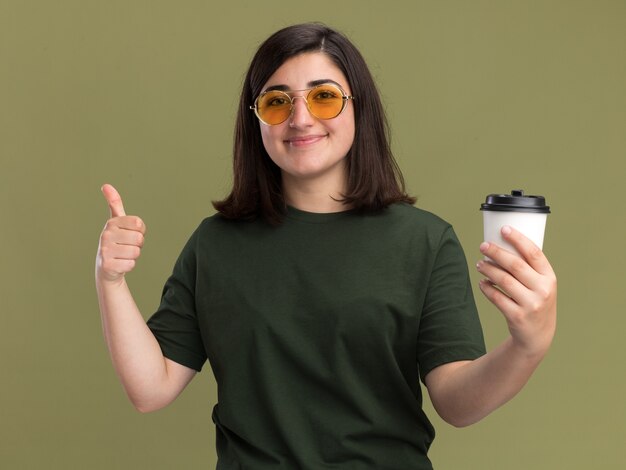 Agradable joven bastante caucásica en gafas de sol pulgar hacia arriba y sostiene un vaso de papel en verde oliva