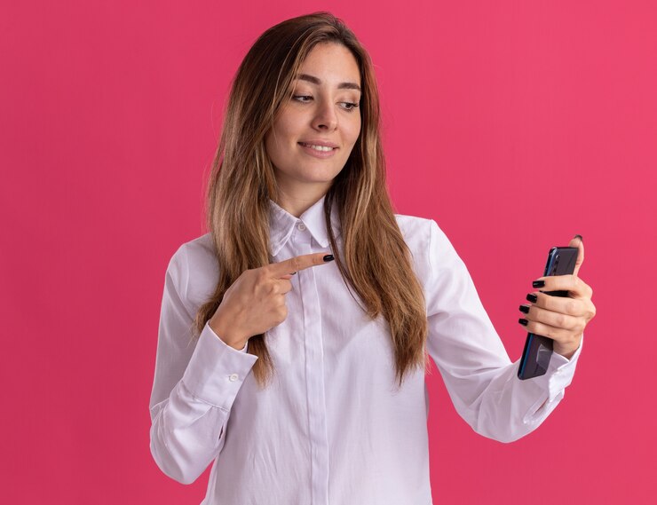 Foto gratuito agradable joven bastante caucásica mira y apunta al teléfono aislado en la pared rosa con espacio de copia