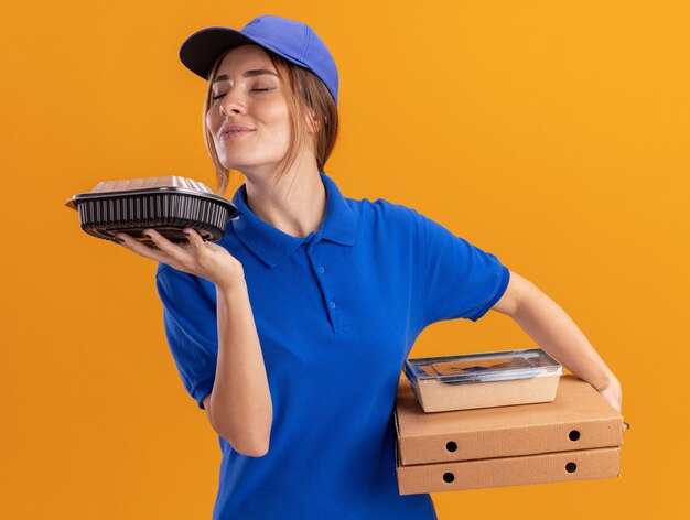 Agradable joven bonita repartidora en uniforme sostiene y huele contenedores de paquetes de alimentos de papel en cajas de pizza en naranja