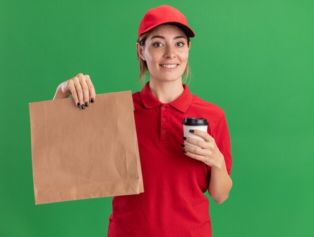 Agradable joven bonita repartidora en uniforme sostiene el paquete de papel y la taza de papel en verde