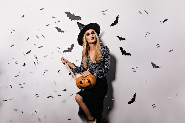 Agradable joven bruja posando con murciélagos en la pared. Chica vampiro bastante rubia con calabaza de halloween.