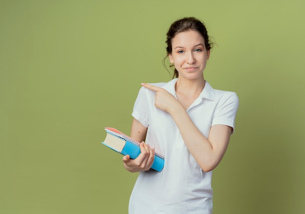 Agradable joven estudiante bastante sosteniendo el libro y el bloc de notas apuntando al lado aislado sobre fondo verde oliva con espacio de copia