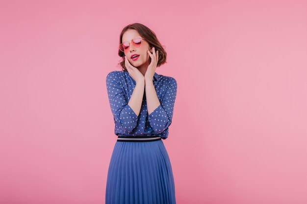 Agradable joven en falda larga azul posando con los ojos cerrados. Jocund modelo femenino rizado en gafas de sol de pie en la pared rosa.