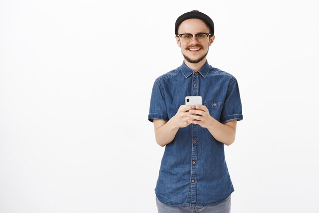 Agradable joven guapo con barba y bigote en gafas y gorro negro con smartphone mirando con encantadora sonrisa feliz