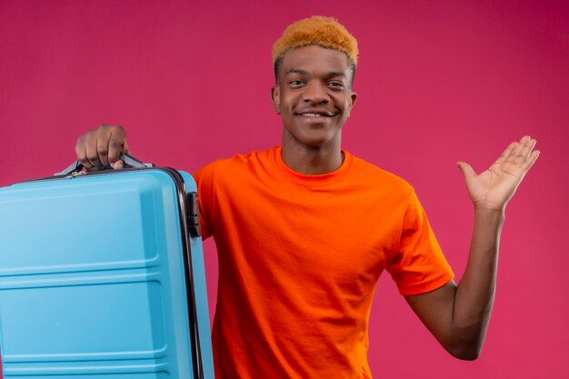 Agradable joven guapo vistiendo camiseta naranja con maleta de viaje sonriendo feliz y positivo con el brazo levantado de pie sobre la pared rosa