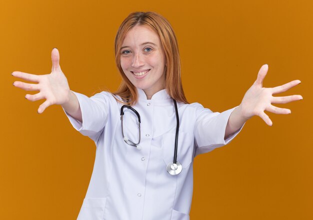 Agradable joven médico de jengibre vistiendo bata médica y estetoscopio a los invitados a la reunión con los brazos abiertos haciendo un gesto de bienvenida