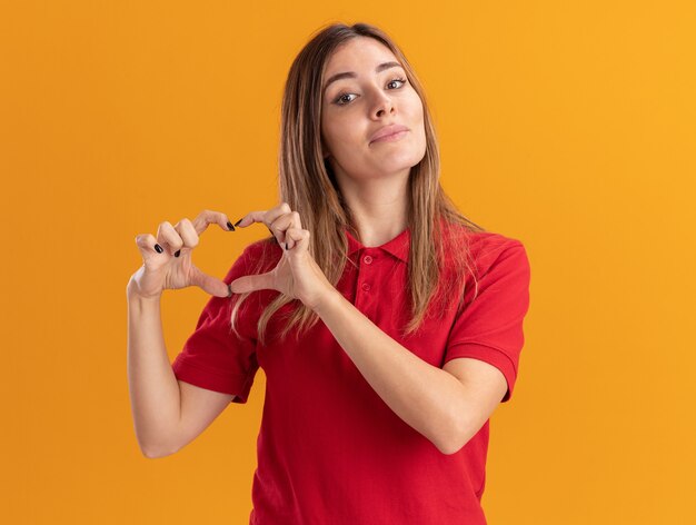 Agradable joven mujer bonita gestos signo de la mano del corazón aislado en la pared naranja