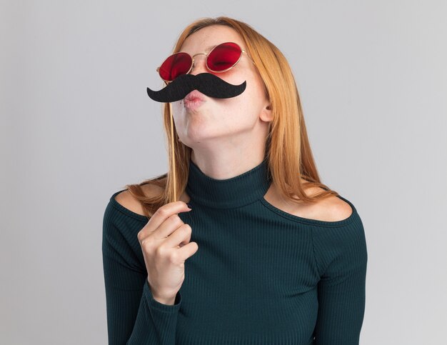 Agradable joven pelirroja pelirroja con pecas en gafas de sol sostiene bigote falso en palo de pie con los ojos cerrados en blanco