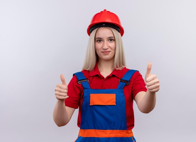 Agradable joven rubia ingeniero constructor chica en uniforme mostrando los pulgares hacia arriba en el espacio en blanco aislado con espacio de copia