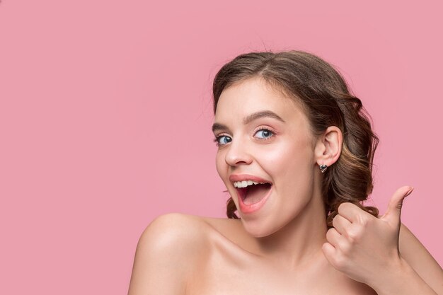 Agradable joven sonriente con cabello largo y sedoso ondulado, maquillaje natural con la mano cerca de la barbilla aislada en la pared rosa. Modelo de piel fresca y brillante y maquillaje natural. Emociones de la gente