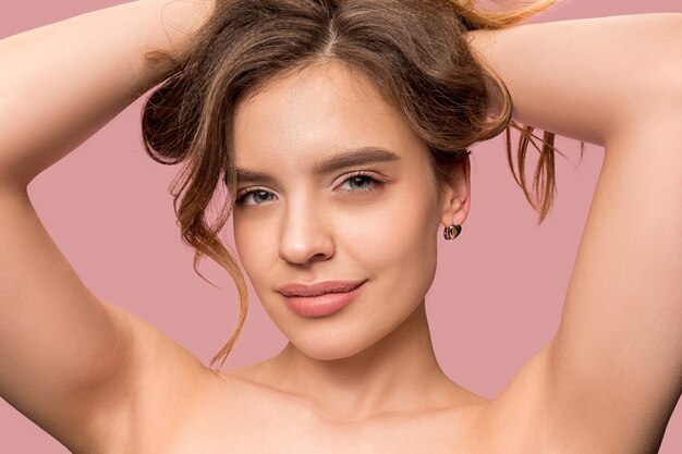 Agradable joven sonriente con cabello largo y sedoso ondulado, maquillaje natural con la mano cerca de la barbilla aislada en rosa