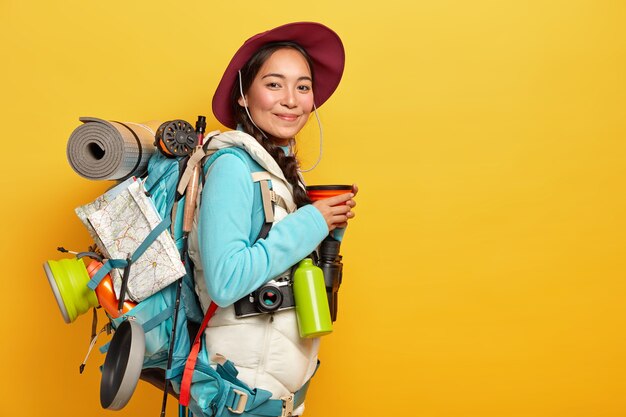 Agradable joven viajera asiática se detiene en su camino para tomar un café, usa sombrero y atuendo informal, posa con mochila, tiene un largo viaje, explora nuevos lugares, le gusta viajar