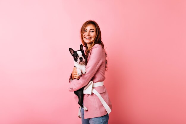 Agradable mujer caucásica en traje rosa abrazando cachorro. Glamorosa chica blanca con bulldog francés con sonrisa.