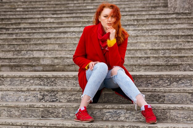 Una agradable mujer joven con el pelo rojo con un abrigo rojo y jeans está sentada en los escalones afuera