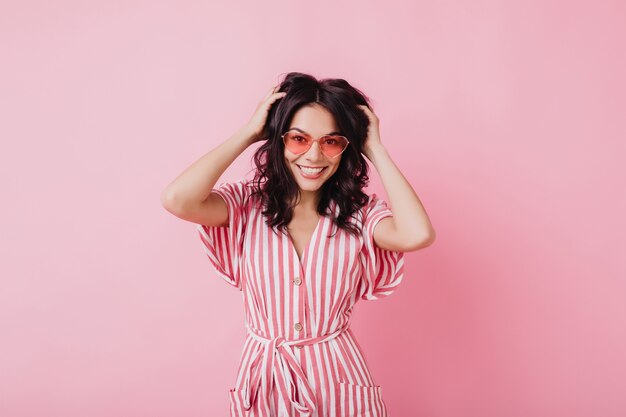 Agradable mujer latina en gafas de sol de moda tocando su cabello oscuro. Foto interior de juguetona chica morena aislada