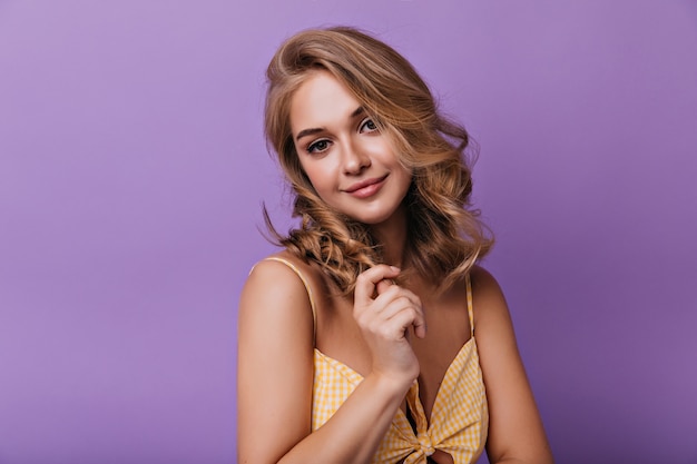 Agradable mujer rubia posando en brillante con una sonrisa suave. Retrato de dama elegante de pie delante de la pared púrpura.