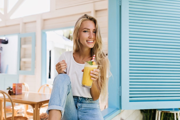 Agradable mujer rubia sentada en la cafetería con un vaso de jugo. Increíble dama bronceada en camiseta blanca riendo mientras posa con cóctel.