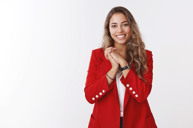 Agradecida y complacida joven hermosa europea de pelo rizado, vestida con una chaqueta roja presionando las palmas juntas agradecidas, apreciando el lindo gesto romántico, sonriendo recibiendo felicitaciones de cumpleaños