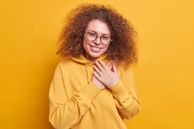 Agradecida y complacida joven de pelo rizado toma las manos cerca del corazón sonríe amablemente dice gracias aprecia ayuda vestida con sudadera aislada sobre pared amarilla. Concepto de lenguaje corporal