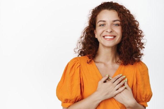 Agradecido. Retrato de mujer sonriente feliz con pelo rojo rizado, tomarse de las manos en el pecho agradecido, decir gracias, apreciar algo, esperanzado por algo en blanco