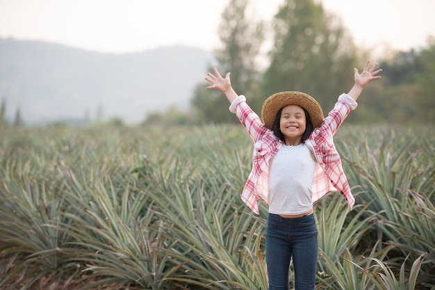 La agricultora asiática ve el crecimiento de la piña en la granja, la muchacha joven bonita del granjero de pie en tierras de cultivo con los brazos levantados hacia la alegría eufórica felicidad.