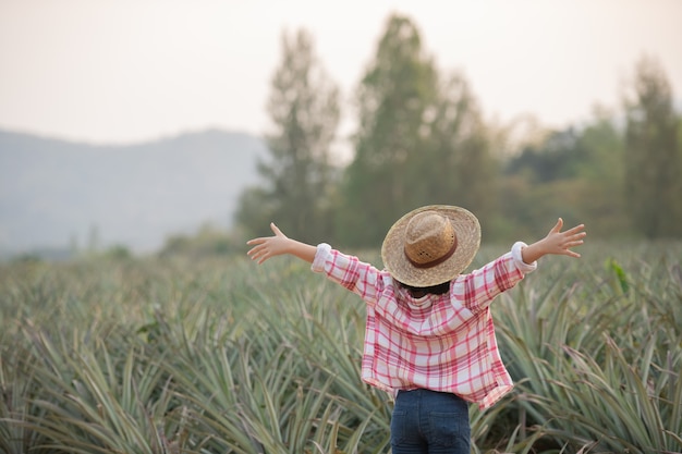 Foto gratuita la agricultora asiática ve el crecimiento de la piña en la granja, la muchacha joven bonita del granjero de pie en tierras de cultivo con los brazos levantados hacia la alegría eufórica felicidad.