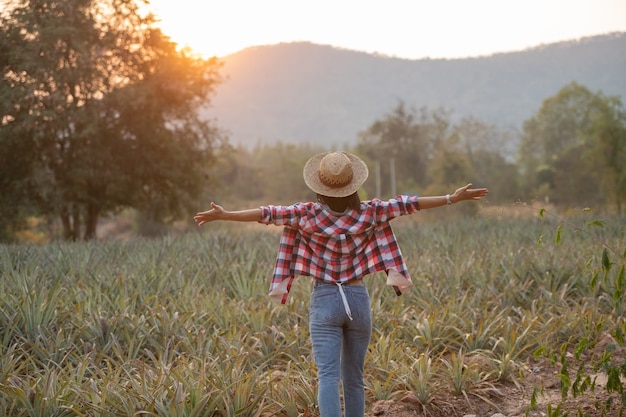 La agricultora asiática ve el crecimiento de la piña en la granja, Mujer joven bonita del granjero de pie en tierras de cultivo con los brazos levantados hacia la alegría eufórica alegría.