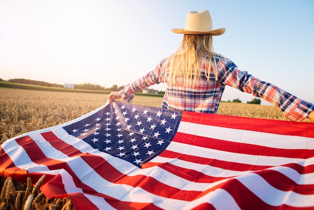 La agricultora estadounidense en ropa casual con los brazos abiertos sosteniendo la bandera de Estados Unidos en el campo de trigo