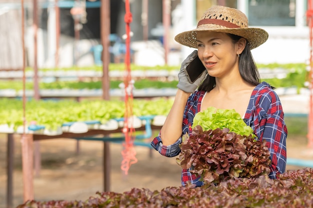 Agricultores asiáticos que trabajan con el móvil en la granja hidropónica de verduras con felicidad. Retrato de mujer agricultora que controla la calidad de la ensalada verde con una sonrisa en la granja de la casa verde.