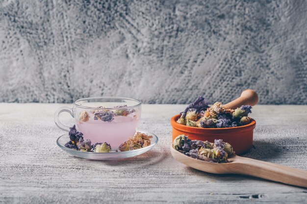 Agua de color púrpura en una taza con té de flores secas en un tazón y cuchara vista lateral sobre un fondo blanco de madera y gris. espacio libre para su texto