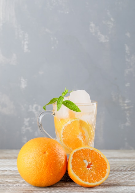 Agua de desintoxicación helada con naranjas, menta en una taza en la pared de madera y yeso, vista lateral.