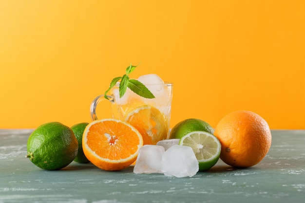 Agua de desintoxicación helada en una taza con naranjas, menta, limas, vista lateral sobre yeso y fondo amarillo