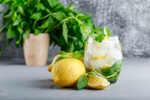 Agua de desintoxicación helada en un vaso con limones y vista lateral de menta sobre superficie gris y grunge