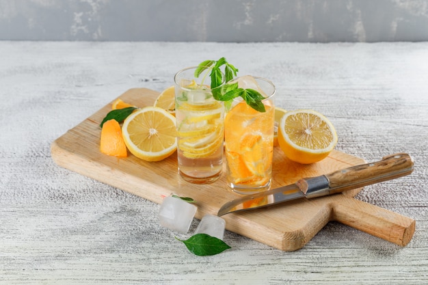 Agua de desintoxicación helada en vidrio con naranjas, limones, menta, cuchillo, tabla de cortar, ángulo de visión alto sobre fondo sucio y yeso