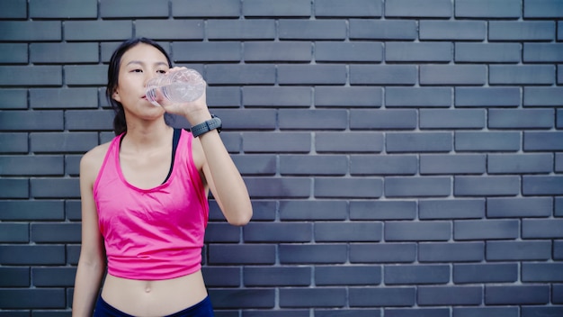 Agua potable de la mujer asiática joven hermosa sana del corredor porque se siente cansada después de correr en la calle