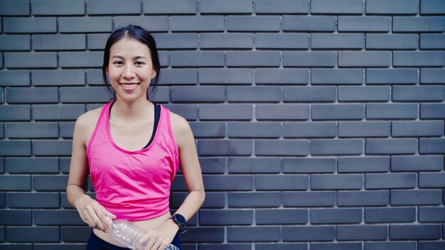 Agua potable de la mujer asiática joven hermosa sana del corredor porque se siente cansada después de correr en la calle