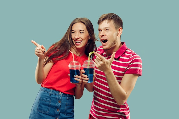 Foto gratuita aislado retrato de medio cuerpo de la hermosa joven pareja. mujer y hombre de pie con bebidas sonriendo y apuntando hacia arriba. expresión facial, verano, concepto de fin de semana. colores de moda.