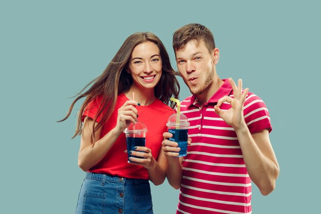 Aislado retrato de medio cuerpo de la hermosa joven pareja. Mujer y hombre de pie con bebidas sonriendo y firmando OK. Expresión facial, verano, concepto de fin de semana. Colores de moda.