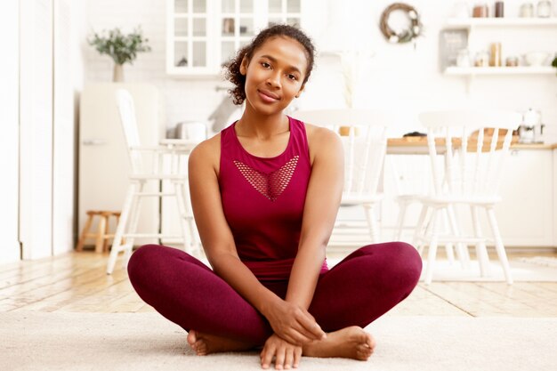 Ajuste lindo joven mujer de piel oscura en ropa deportiva de moda sentado descalzo sobre la estera con las piernas dobladas. Chica negra activa sana relajante en padmasana después de la práctica de yoga